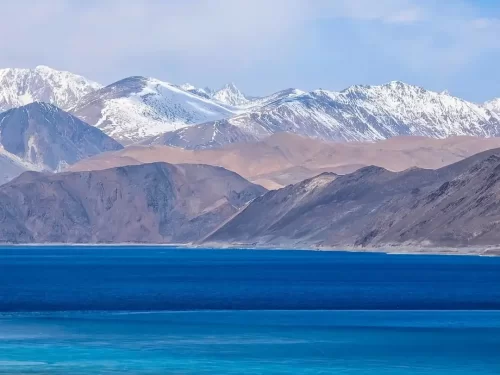 Ladakh Pangong Tso Lake deep blue turquoise brackish water snow capped rugged Himalayan mountains terracotta brown barren slopes scattered clouds pale blue sky high altitude scenic landscape adventure tour package