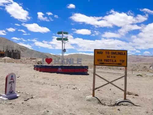 Large red I Love Hanele signpost yellow road sign Way to Umlingla 63 km via Photila Top green directional poles barren Ladakh rocky terrain partly cloudy blue sky, perfect high-altitude route Ladakh adventure package.