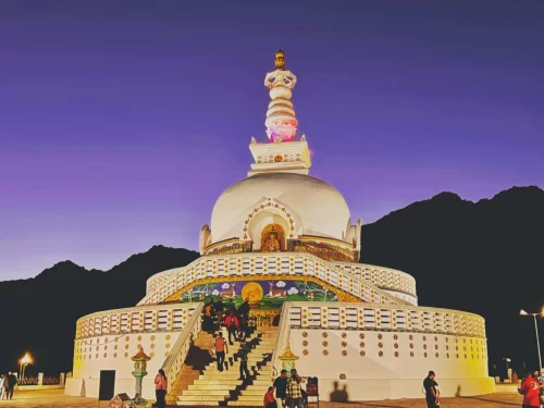 Shanti Stupa Leh evening with tourists ascending staircase illuminated pink purple lights white dome mountains dusk sky, perfect Ladakh spiritual tour package.