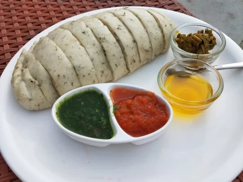 A plate of sliced siddu, a steamed Himachali bread, served with green chutney, red chutney, ghee, and a small bowl of spicy pickle.