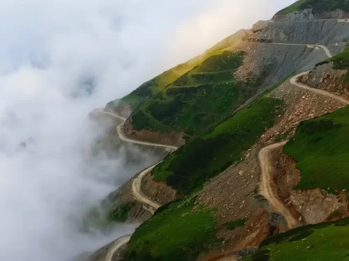 Sinthan Top misty mountain passes with winding roads & sun rays piercing clouds over green slopes, perfect Kashmir adventure tour packages.