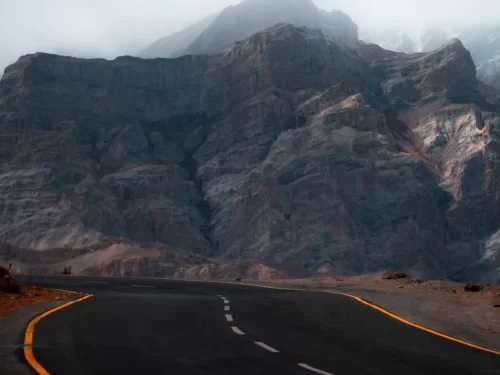 Curving Srinagar Leh Highway asphalt road through dramatic rocky canyon near Lamayuru Ladakh during foggy overcast day, featuring towering gray mountains distant peaks travelers, perfect adventurous drive experience with Srinagar Leh Highway tour package.