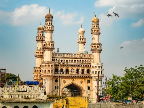 Front view of Charminar at Hyderabad featuring four minarets intricate arches birds flying under partly cloudy skies, ideal for Telangana tour packages.
