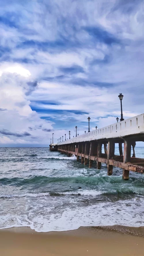 Scenic view of the Promenade Pier in Puducherry, Tamil Nadu, with waves crashing along the sandy shore under a dramatic sky, a popular seaside attraction featured in Tamil Nadu tour packages.