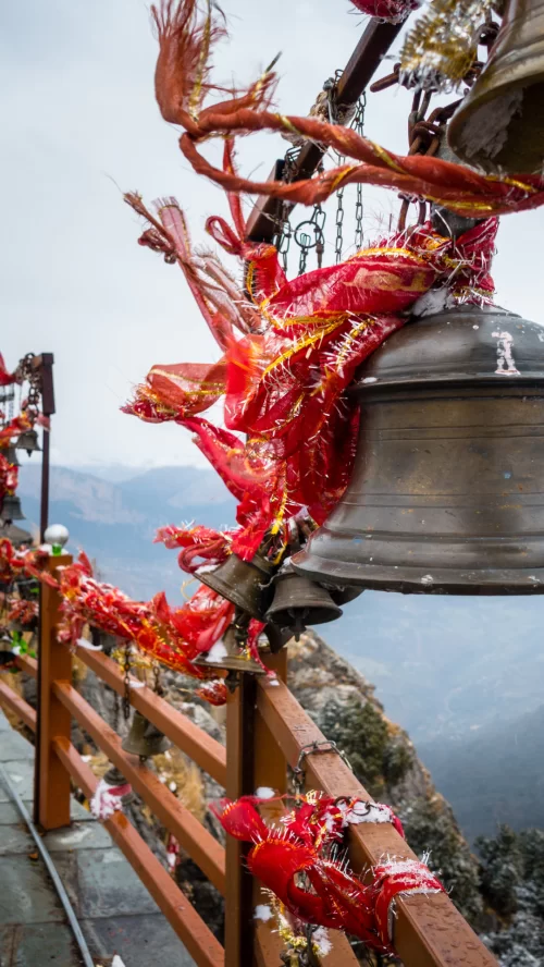 Red temple bells adorned with marigold garlands and red threads at Kartik Swami Temple viewpoint, overlooking misty Uttarakhand mountains perfect for spiritual Garhwal tour packages.