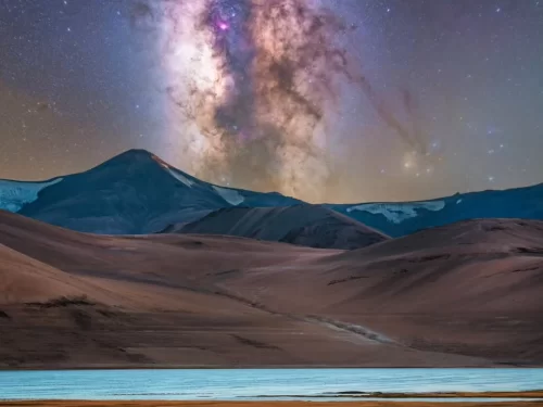 Starry Milky Way galaxy arching over snow-dusted dark rugged mountains turquoise lake foreground reddish brown arid hills Tso Kar Wetland Complex Ladakh India at night, perfect astrophotography stargazing camping adventure with Leh Ladakh tour package. 