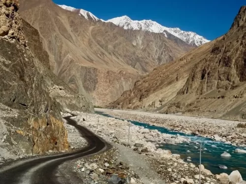 Scenic winding road along Shyok River to Turtuk village Nubra Valley Ladakh during clear blue sky, featuring snow-capped Karakoram peaks, barren gorges, perfect adventure Turtuk tour package. 